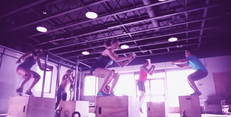 A group of people in athletic wear perform box jumps in a gym with a high ceiling. The scene is energetic, conveying a sense of determination.