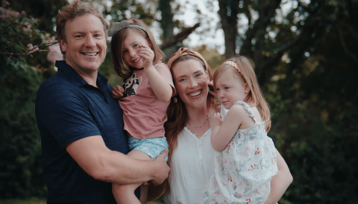 A smiling family of four outdoors, with parents holding two young children, both in dresses. The background features lush green trees, suggesting a joyful, sunny day.