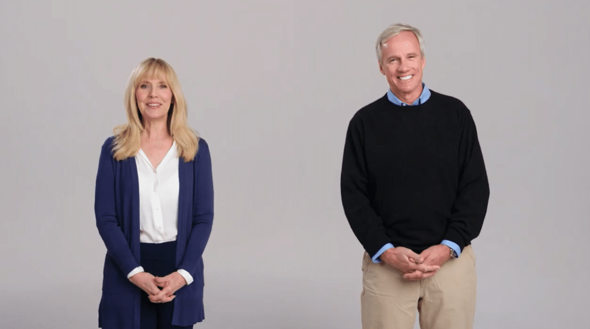 A smiling woman in a blue cardigan and a man in a black sweater stand against a gray background, conveying a friendly and approachable vibe.