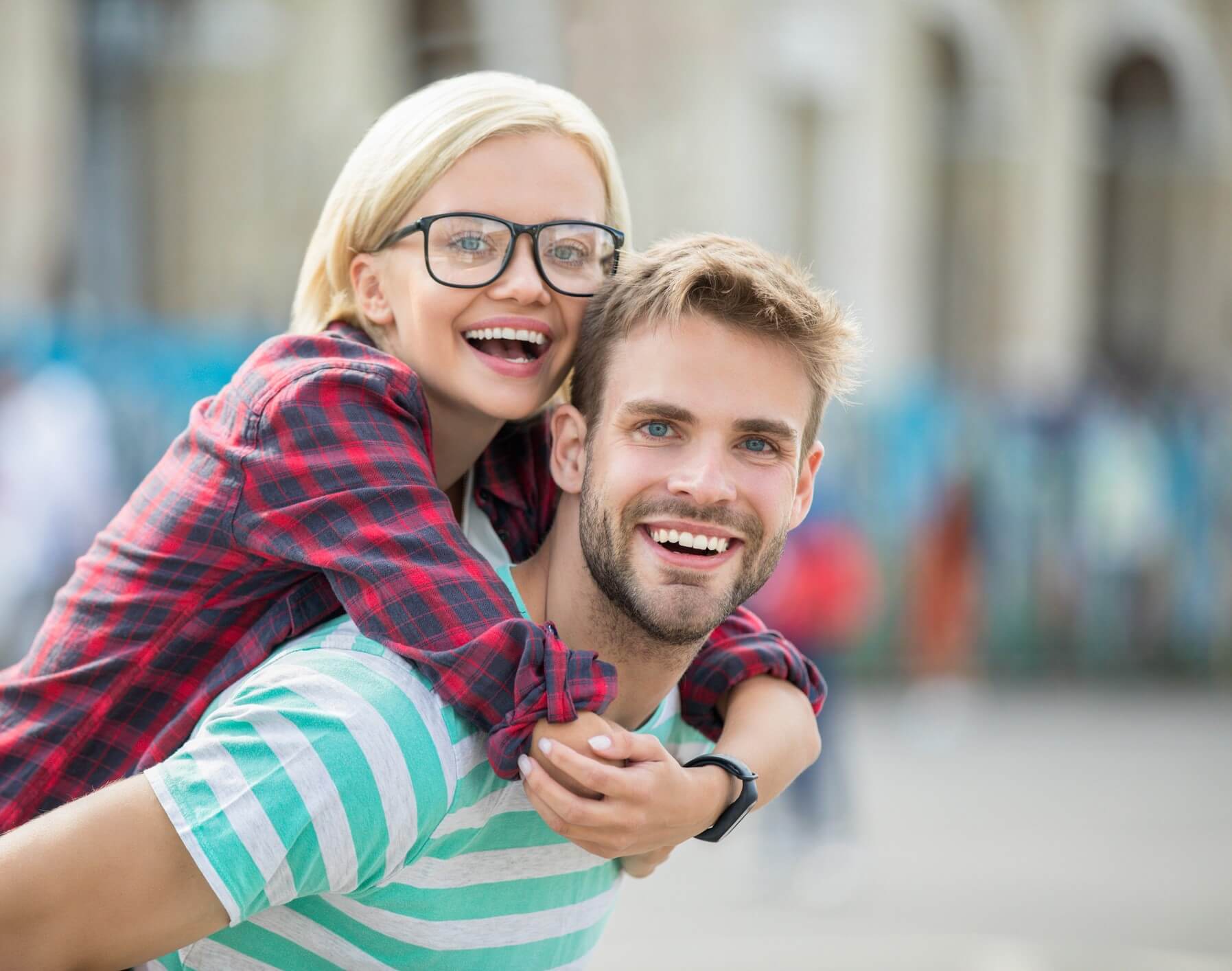 A cheerful woman in glasses playfully piggybacks on a smiling man. She wears a red plaid shirt; he is in a green striped tee. Both exude happiness in a blurred urban setting.