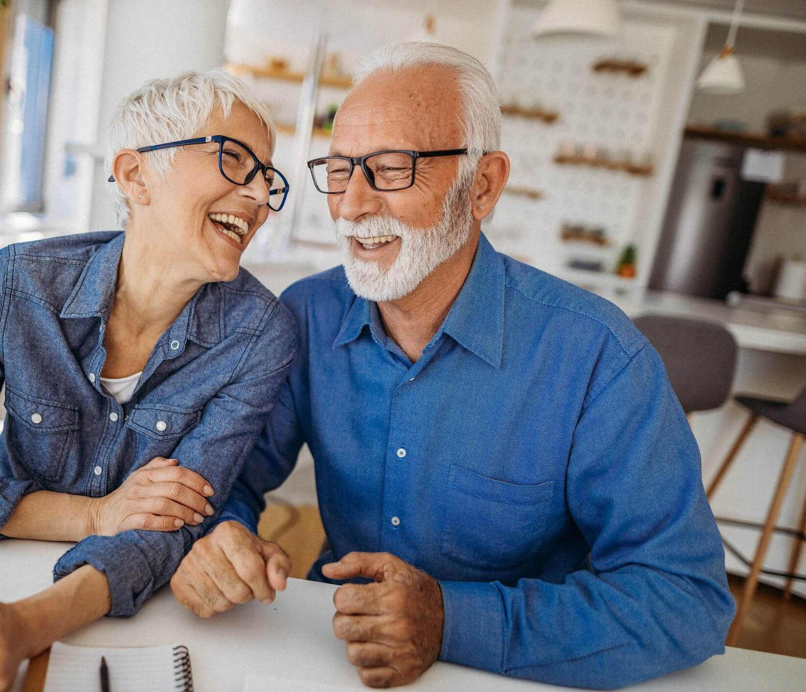 Elderly couple sits at a table in a bright kitchen, both wearing glasses and denim shirts, smiling and laughing warmly, exuding joy and companionship.