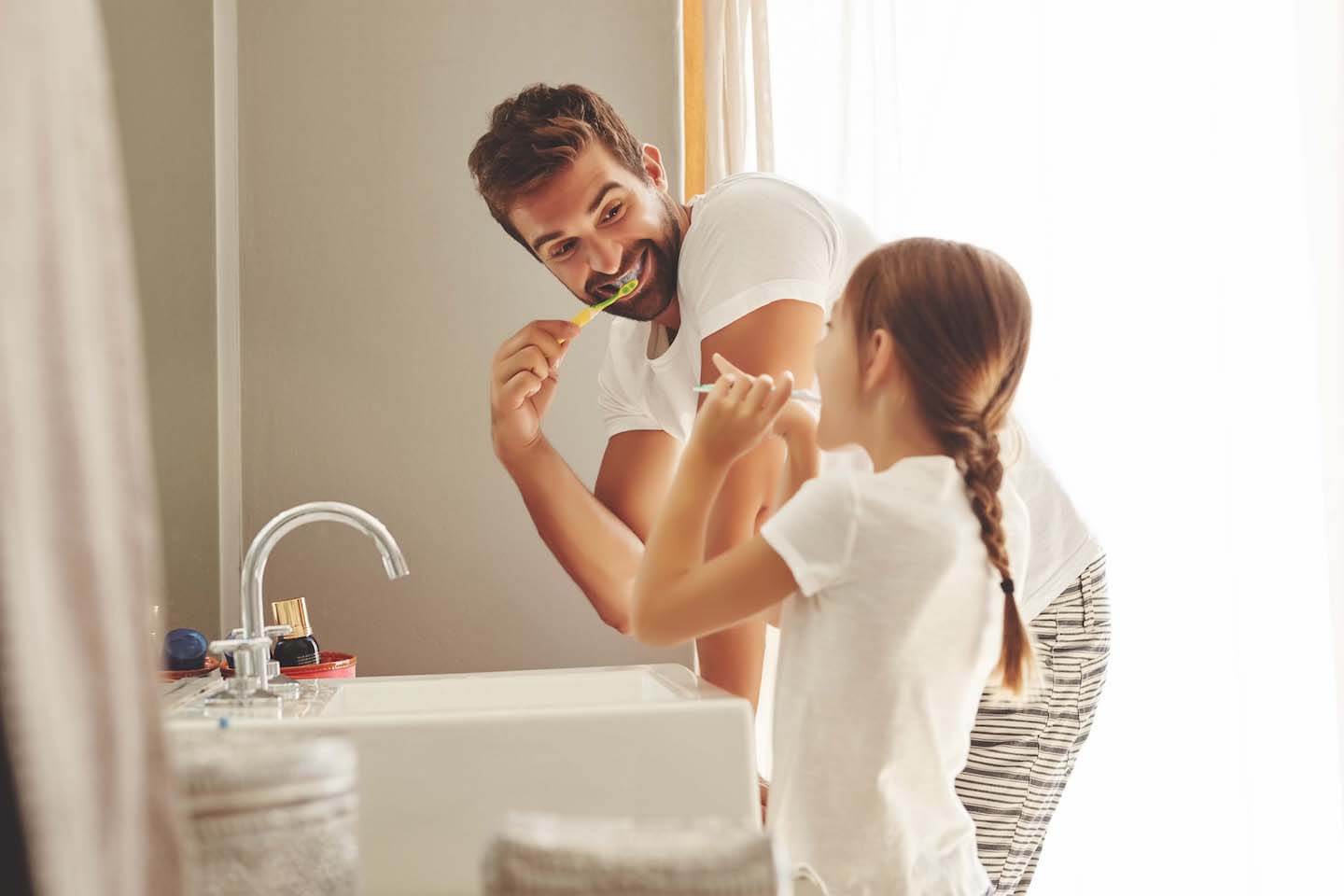 Father and daughter joyfully brush teeth together in a bright bathroom. Both smile in matching white shirts, capturing a moment of playful bonding.