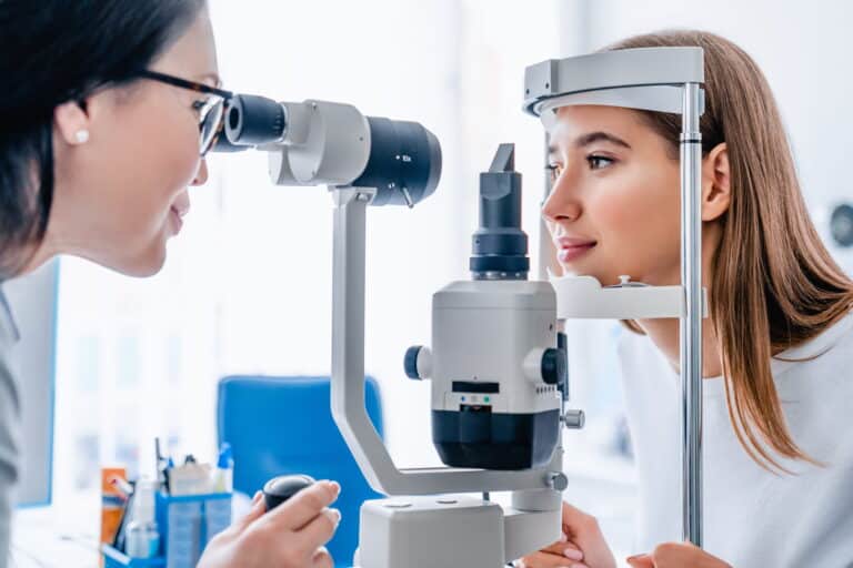 An optometrist examines a patient's eyes using a slit lamp in a bright clinic. The atmosphere is professional and focused on eye care.