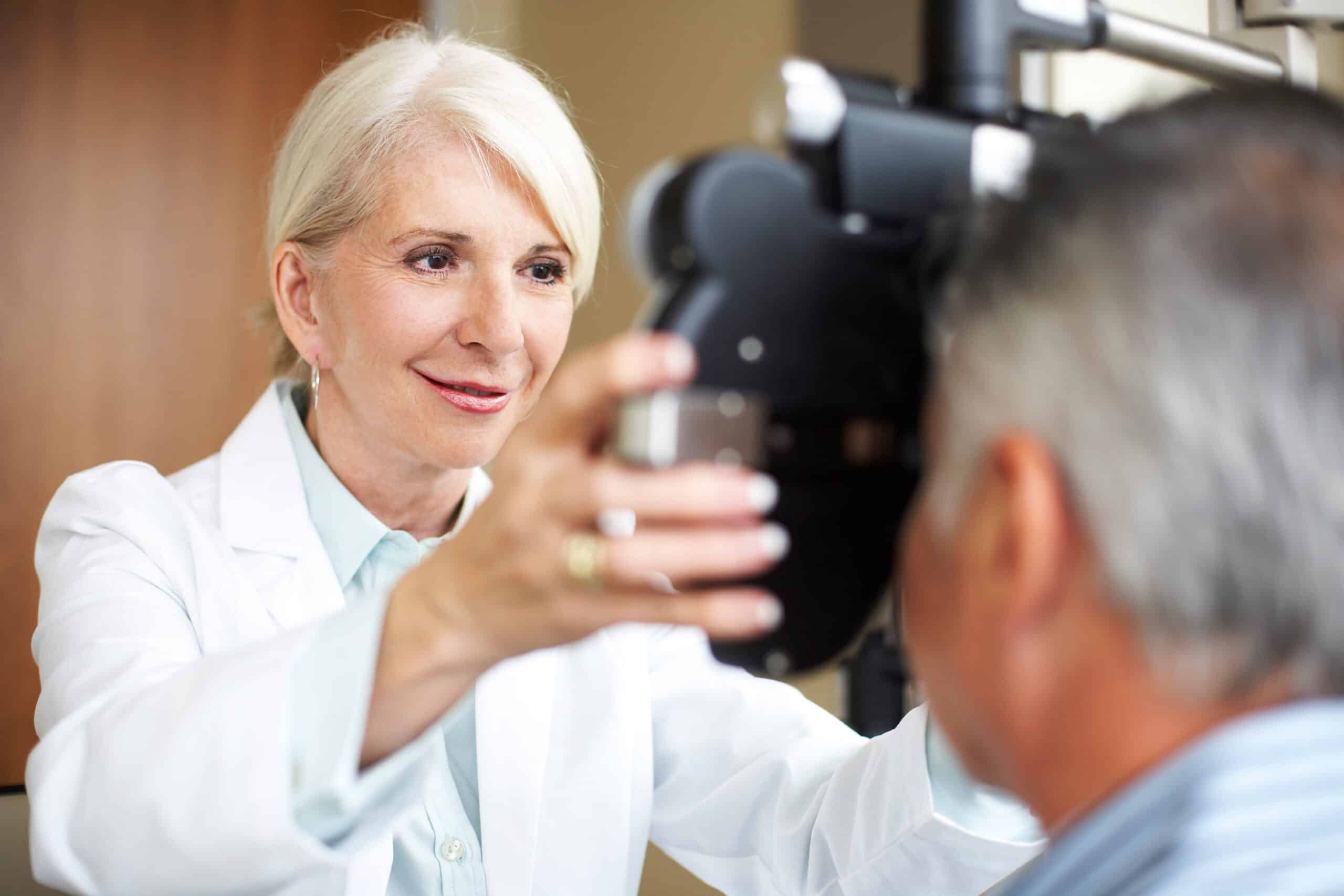 An elderly woman in a white coat performs an eye exam on a man using optometry equipment. Her expression is focused and attentive.