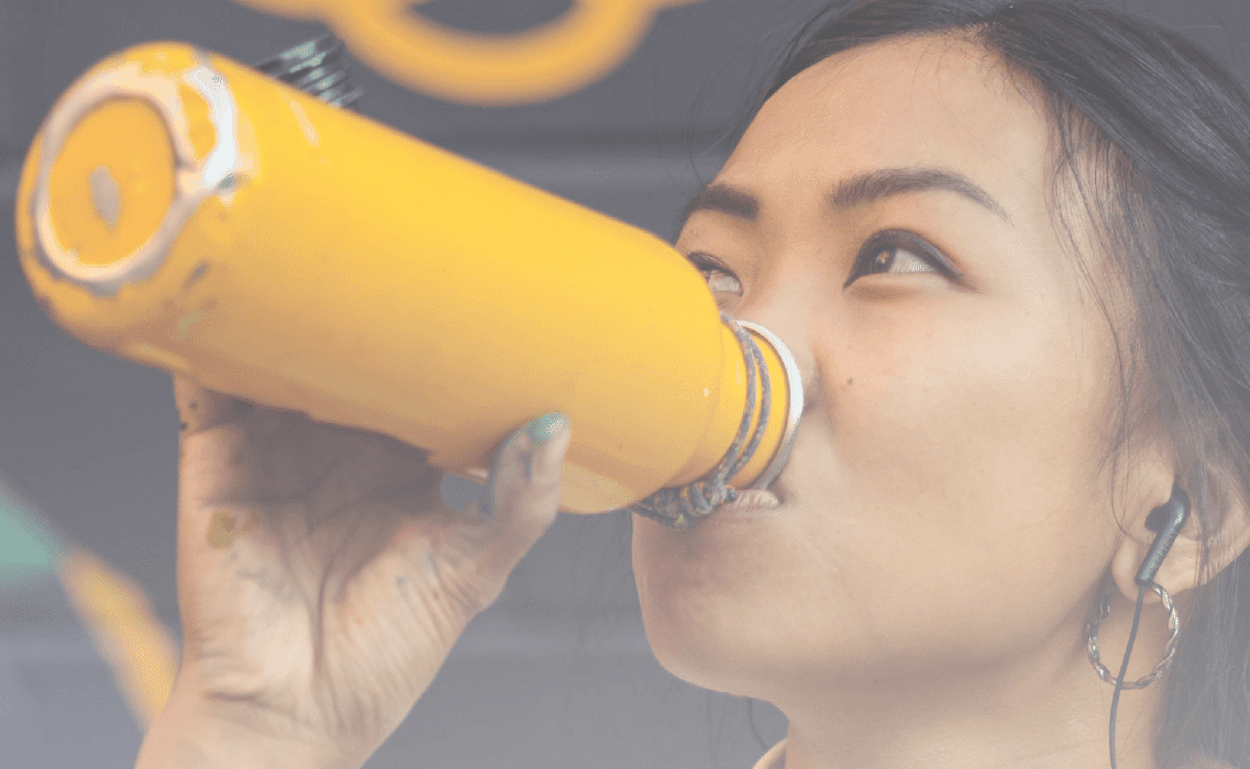 A woman drinks from a yellow bottle while wearing earbuds, with a colorful graffiti-covered wall in the background. The scene conveys vibrancy and energy.