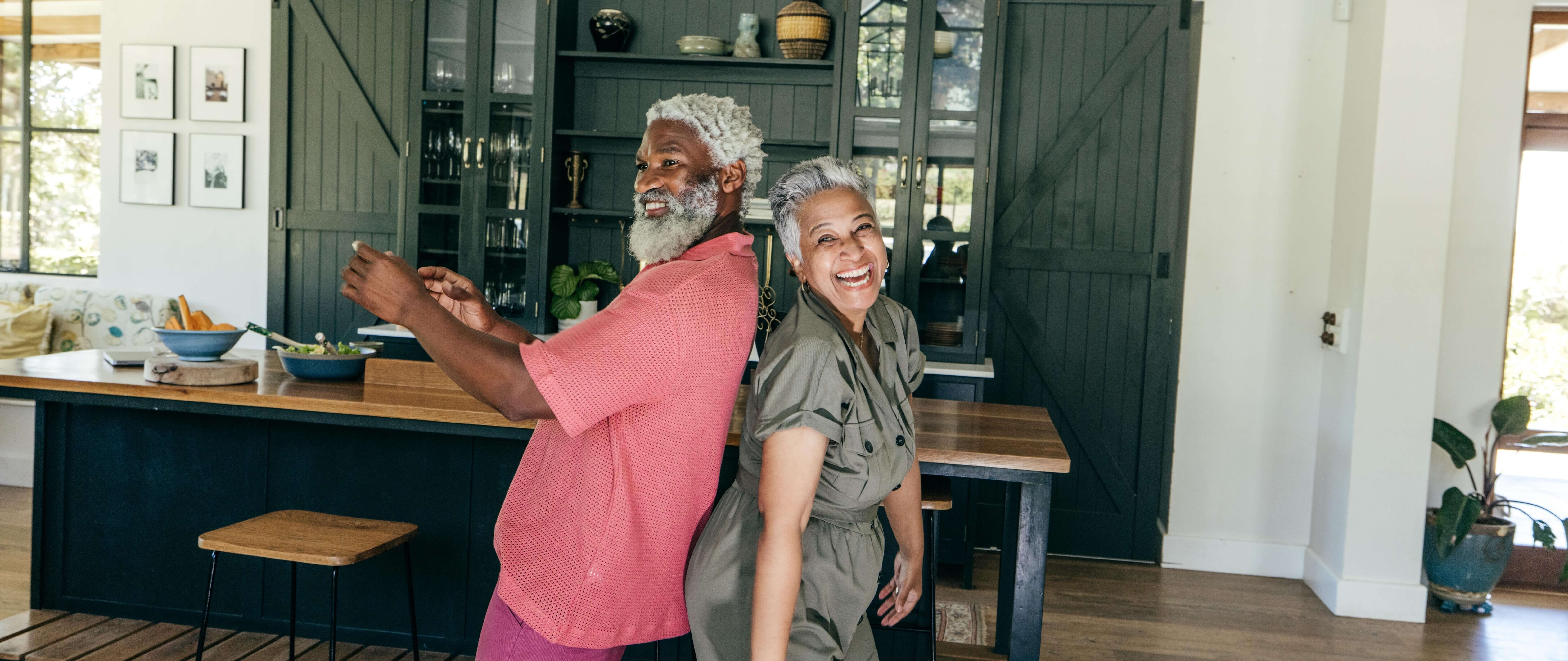 A joyful older couple dances back-to-back in a modern kitchen, smiling widely. Wooden cabinets and houseplants create a warm, inviting atmosphere.