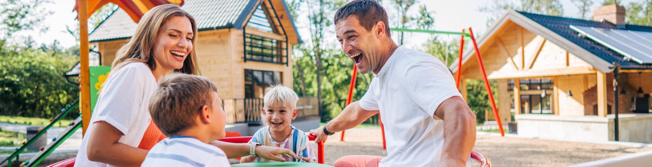 A joyful family of four plays on a merry-go-round in a sunlit playground, surrounded by modern wooden houses and lush trees, conveying a sense of happiness.