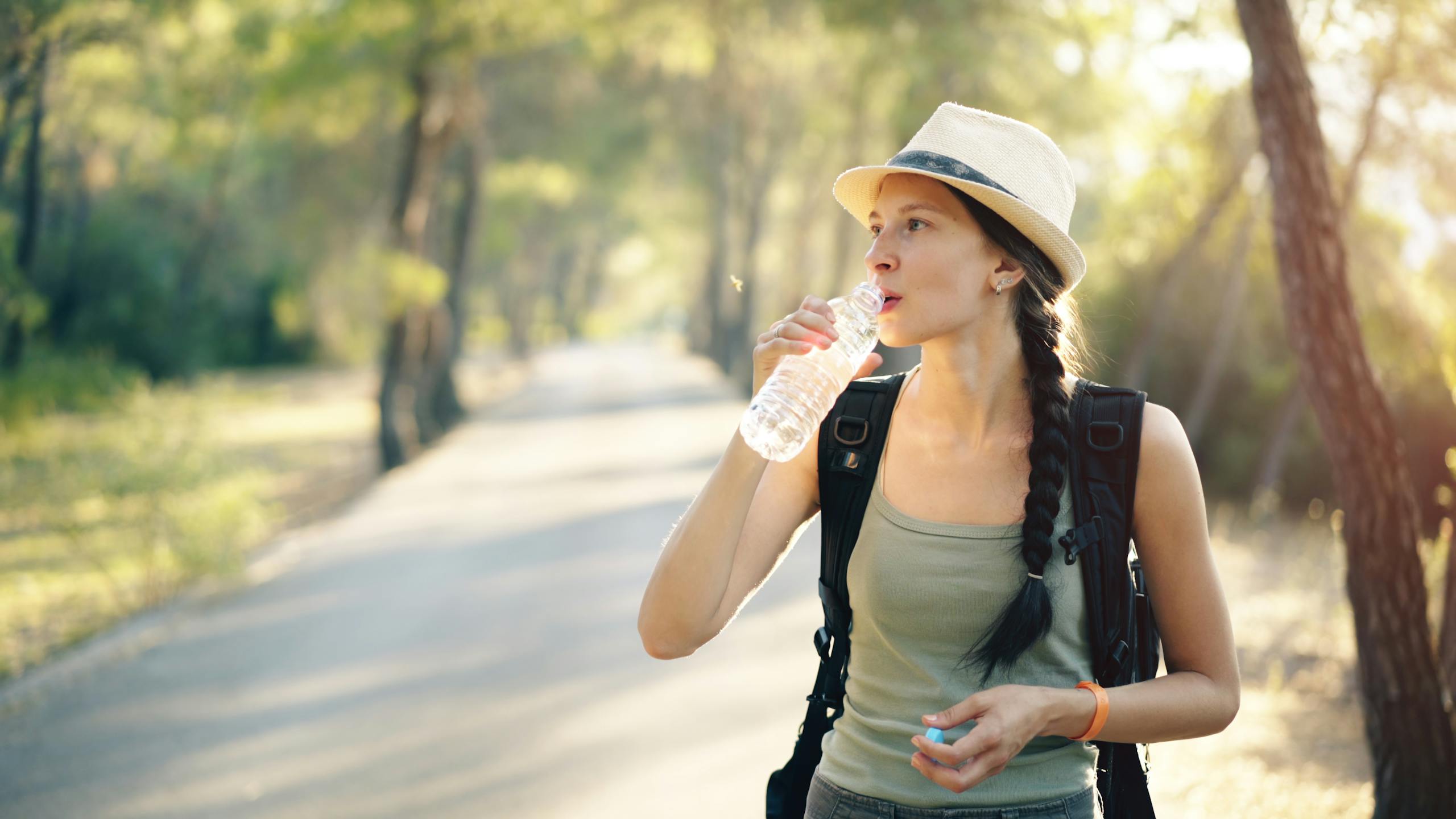 Young woman hiker in tank top and hat drinking water on a forest trail.